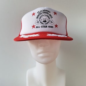 May include: A red and white trucker hat with the text "12th Annual Winter Tribal Days Dakota Ojibway Tribal Council All-Star 1986." The hat has a red brim and a white front panel with red stars.