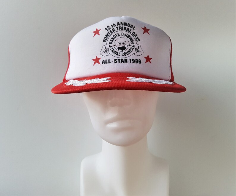 May include: A red and white trucker hat with the text "12th Annual Winter Tribal Days Dakota Ojibway Tribal Council All-Star 1986." The hat has a red brim and a white front panel with red stars.