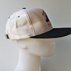 Op de afbeelding: Een beige en zwarte baseballcap met een platte klep. De cap heeft een geborduurd ontwerp met de tekst "Truck & Equipment" en "FINISH". De klep is zwart en de kroon beige, met een verstelbare band aan de achterkant.