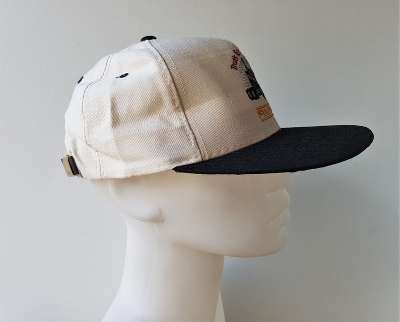 Op de afbeelding: Een beige en zwarte baseballcap met een platte klep. De cap heeft een geborduurd ontwerp met de tekst "Truck & Equipment" en "FINISH". De klep is zwart en de kroon beige, met een verstelbare band aan de achterkant.