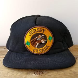 May include: A black baseball cap featuring a circular embroidered patch on the front. The patch has a yellow background with the text "JERSEY CANADA" in brown, a brown and white cow illustration, and two small green maple leaf icons on the sides.