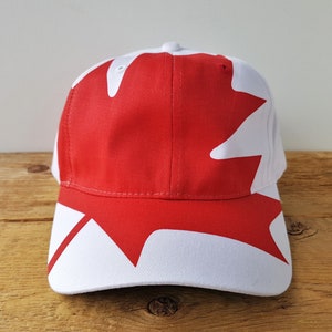 May include: A red and white baseball cap with a maple leaf design. The cap has a curved peak and a structured crown. The maple leaf design is a prominent feature on the front of the cap.