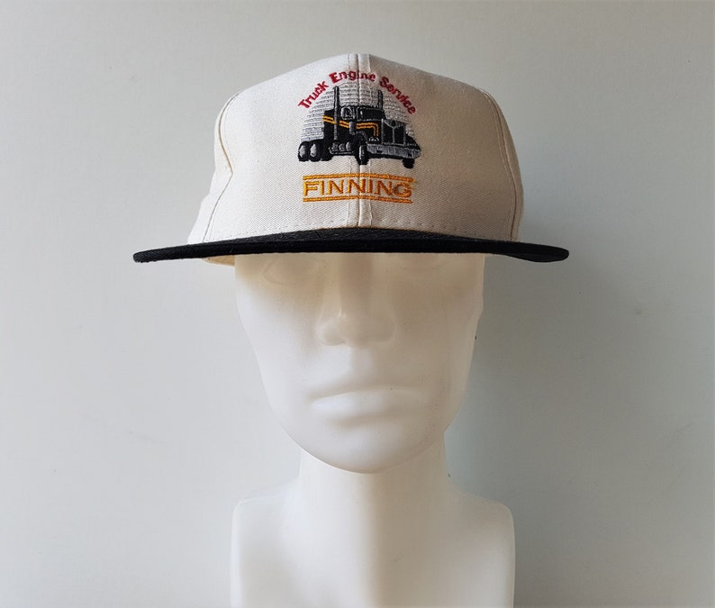 Op de afbeelding: Beige en zwarte baseballcap met de tekst "Truck Engine Service" en de naam "FINNING" geborduurd in oranje. De cap heeft een afbeelding van een vrachtwagen.
