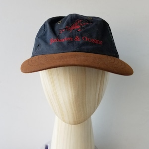 May include: A navy blue baseball cap with a brown peak. The cap has red embroidered text and a fish design, reading "Fisheries & Oceans". The hat is displayed on a wooden mannequin head.