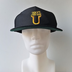 May include: Black baseball cap with a flat peak and a green underside. The cap has the embroidered text "UKROP'S OWN BEEF" in gold, with a gold "U" below. The cap is displayed on a white mannequin head.