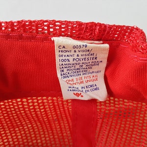 May include: A red trucker hat with a mesh back and a solid front. The hat has a tag with text including "CA. 00579" and "ONE SIZE FITS ALL". The hat is made in Korea.
