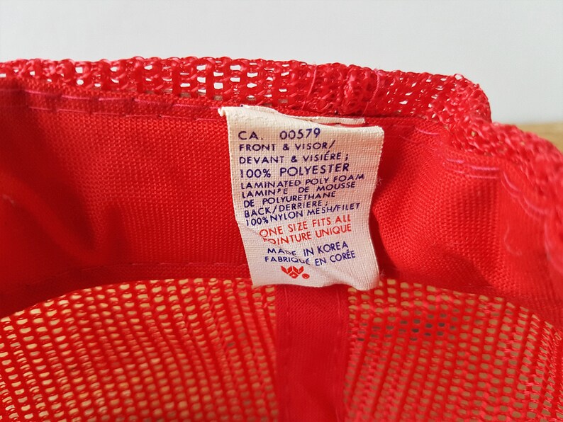 May include: A red trucker hat with a mesh back and a solid front. The hat has a tag with text including "CA. 00579" and "ONE SIZE FITS ALL". The hat is made in Korea.