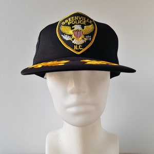 Op de afbeelding: Zwarte truckercap met een gouden geborduurde patch met de tekst "Greenville Police" en een adelaar met een schild en de letters "N.C."