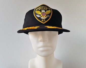May include: Black trucker hat with a gold embroidered patch that says "Greenville Police" and features an eagle with a shield and the letters "N.C."