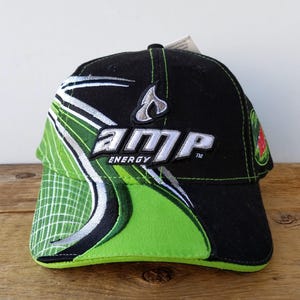 Puede incluir: Gorra de béisbol negra con detalles verdes y blancos. La gorra presenta el logotipo "AMP Energy" en blanco, con un diseño de llama. La visera y los paneles laterales son verdes, con un patrón de rejilla. Una pequeña etiqueta está adjunta.