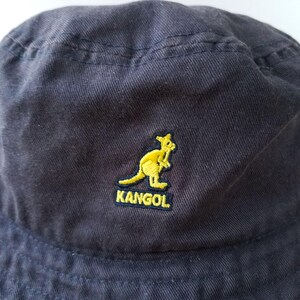 K&ouml;nnte beinhalten: Marineblauer Fischerhut mit gelbem K&auml;nguru-Logo und dem gestickten Wort "KANGOL". Der Hut ist aus weichem, strukturiertem Stoff gefertigt und hat einen l&auml;ssigen Stil.