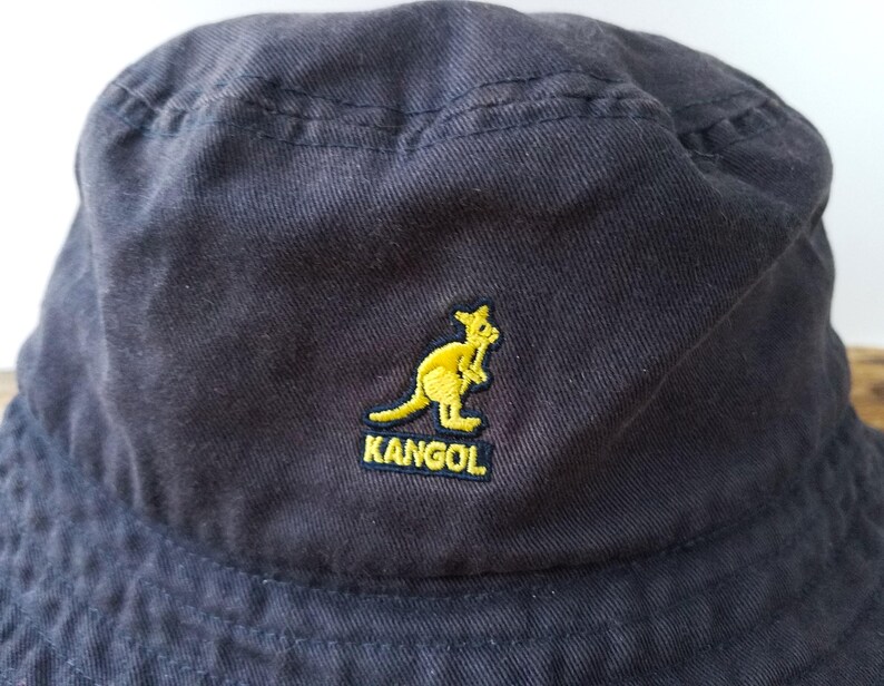 K&ouml;nnte beinhalten: Marineblauer Fischerhut mit gelbem K&auml;nguru-Logo und dem gestickten Wort "KANGOL". Der Hut ist aus weichem, strukturiertem Stoff gefertigt und hat einen l&auml;ssigen Stil.
