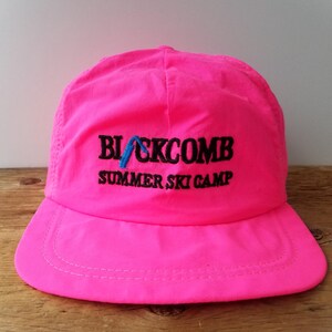 May include: Vibrant pink baseball cap featuring the text "BLACKCOMB SUMMER SKI CAMP" in black and blue embroidery. The cap has a curved peak and a structured crown, suitable for outdoor pursuits.