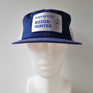 May include: A blue and white trucker hat with the text "Apprentice Bozon Hunter" on a rectangular patch. The hat has a mesh back and a curved brim. The patch features a cartoon character.