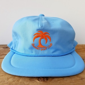 May include: Pale blue golf cap with a flat peak and a rope trim. The cap has an embroidered orange palm tree and the words "Busuky Golf Club". The cap is made of a lightweight material and has a snapback closure.