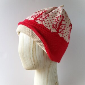 May include: A red, cream, and beige knit hat with a winter tree pattern. The hat is displayed on a wooden mannequin head. The hat has a red brim and a cream-colored interior. The top of the hat has a small, cream-colored point.