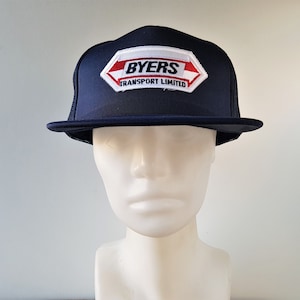 May include: A navy blue trucker hat with a white mesh back. The hat has a white patch with red lettering that says "BYERS TRANSPORT LIMITED".