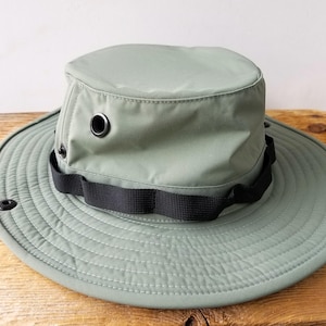 May include: A sage green sun hat with a wide brim and a black band. The hat has a structured crown with two eyelets and a black strap. The brim is stitched with concentric circles. This hat is designed for outdoor activities.