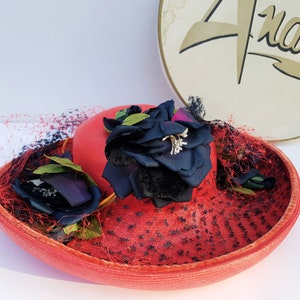 May include: A red straw hat with a wide brim, adorned with a large navy blue flower and smaller dark blue accents. The hat features a red netting and a gold-colored band. The word "Andre" is visible in the background.