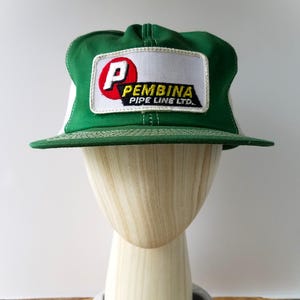 May include: Green and white trucker hat with a rectangular patch. The patch features a red "P" inside a white circle, with the text "PEMBINA PIPE LINE LTD." in black and yellow. The hat is displayed on a wooden mannequin head.