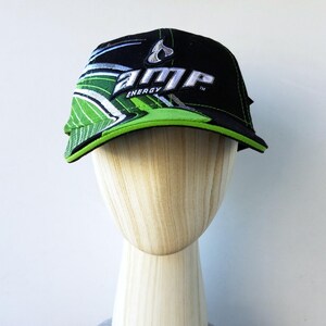 Puede incluir: Gorra de béisbol negra con el texto "AMP ENERGY" en blanco y verde. La gorra tiene una visera curva con detalles en verde y negro. El sombrero se exhibe en una cabeza de maniquí de madera.