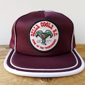 May include: A burgundy trucker hat with a white brim and trim. The hat features a circular patch with the text "BELLA COOLA B.C. HOME OF THE THUNDERBIRD" and a graphic of a Thunderbird.