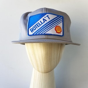 Puede incluir: Gorra de camionero gris con visera plana. La gorra presenta un parche rectangular con el texto "ROTELLA'T" en blanco, un fondo azul y un logotipo de Shell. La gorra se muestra en una cabeza de maniquí de madera.