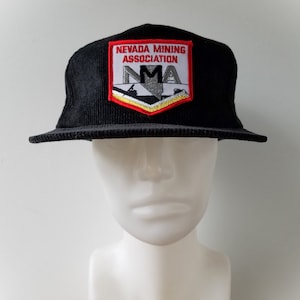 Puede incluir: Gorra de béisbol de pana negra con un parche bordado en rojo, blanco y amarillo que dice "Nevada Mining Association" y "NMA".