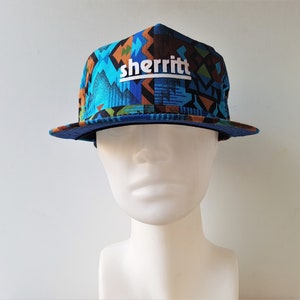 May include: A flat-brimmed hat with a geometric pattern in shades of blue, brown, and orange. The hat features the word "sherritt" in white block letters above a mountain graphic. The brim and underside are solid blue.