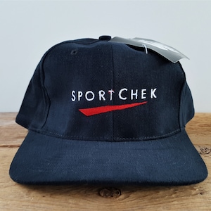 Pode incluir: Um boné de beisebol azul marinho com o texto branco "SPORTCHEK" e um traço vermelho. O boné tem uma aba curva e uma etiqueta anexada. O chapéu está em uma superfície de madeira.