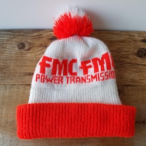 Può includere: Un berretto bianco lavorato a maglia con bordo e pompon arancioni. Il cappello presenta la scritta "FMC FM POWER TRANSMISSION" in lettere arancioni in grassetto. Il berretto è realizzato in morbido materiale a maglia.