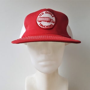 Może przedstawiać: Czerwono-biała czapka trucker z haftowanym czerwonym logo z napisem "Ted Doyle Trucking" i białym ciągnikiem siodłowym.