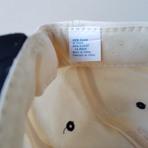 Op de afbeelding: Cr&egrave;mekleurige baseballpet met een marineblauwe rand. De binnenkant toont een label met tekst waaronder "100% Cotton" en "Made in China". De pet heeft een zwart geborduurd detail.