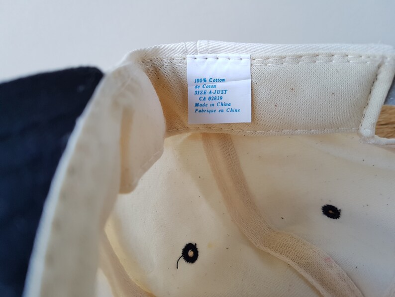 Op de afbeelding: Cr&egrave;mekleurige baseballpet met een marineblauwe rand. De binnenkant toont een label met tekst waaronder "100% Cotton" en "Made in China". De pet heeft een zwart geborduurd detail.