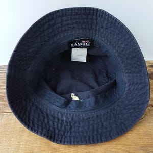 K&ouml;nnte beinhalten: Marineblauer Fischerhut mit dem Markennamen "KANGOL" auf einem Etikett im Inneren. Der Hut hat ein kleines gesticktes Design und ist aus strukturiertem Stoff gefertigt. Der Hut liegt auf einer Holzoberfl&auml;che.