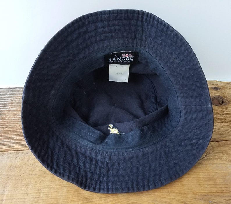K&ouml;nnte beinhalten: Marineblauer Fischerhut mit dem Markennamen "KANGOL" auf einem Etikett im Inneren. Der Hut hat ein kleines gesticktes Design und ist aus strukturiertem Stoff gefertigt. Der Hut liegt auf einer Holzoberfl&auml;che.