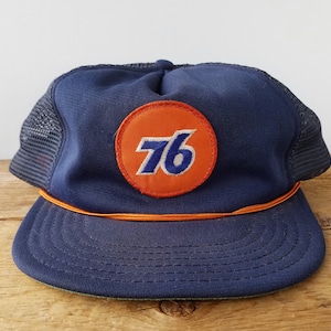 Pode incluir: Boné de caminhoneiro azul marinho com parte traseira em tela e aba curva. A frente apresenta um patch circular laranja com o número "76" em branco e azul. Um cordão laranja adorna a aba.