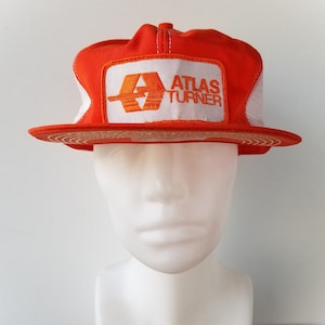 Puede incluir: Gorra de camionero naranja y blanca con un parche blanco que dice "Atlas Turner" en letras naranjas. El parche también presenta un logotipo naranja estilizado.