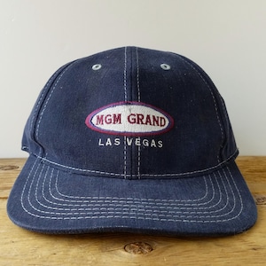 May include: A navy blue baseball cap with white contrast stitching. The front features an embroidered oval patch with a maroon border containing the text MGM GRAND in maroon letters, with the words LAS VEGAS embroidered below in white.