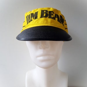 Puede incluir: Gorra con visera amarilla y negra con el texto "JIM BEAM" en letras negras. El sombrero tiene una corona de tela amarilla y una visera negra texturizada. El sombrero se exhibe en una cabeza de maniquí blanca.