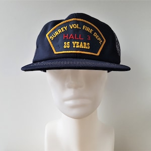 May include: A blue and black trucker hat with a yellow and red embroidered patch that reads "Surrey Vol. Fire Dept. Hall 3 25 Years".