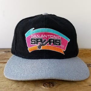 May include: Black and grey San Antonio Spurs baseball cap. The cap features the team's logo in a colourful design with the words "San Antonio Spurs". The brim is grey, and the crown is black.