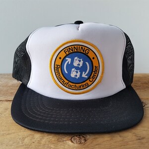 Op de afbeelding: Witte en zwarte truckercap met een ronde gele en blauwe patch waarop "Finning Remanufacturing Center" staat, met een blauwe pijl en twee blauwe tandwielen in de cirkel.