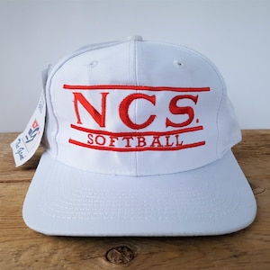 Op de afbeelding: Een witte baseball cap met een platte klep. De cap heeft de rode geborduurde tekst "NCS SOFTBALL" over de voorkant. Een label is aan de zijkant bevestigd. De cap wordt weergegeven op een houten oppervlak.