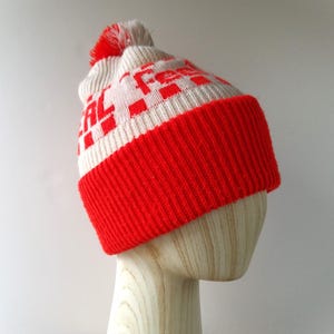 Pode incluir: Um gorro de malha vermelho, branco e creme com um pompom. O chapéu apresenta uma aba canelada vermelha e um padrão xadrez branco e vermelho na coroa. O chapéu é exibido em uma cabeça de manequim de madeira.