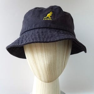 K&ouml;nnte beinhalten: Dunkelblauer Fischerhut mit gelbem K&auml;nguru-Logo. Der Hut hat ein leicht abgenutztes Aussehen und eine breite Krempe. Der Hut wird auf einem Schaufensterkopf pr&auml;sentiert. Durchmesser ca. 20 cm.