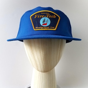 May include: A blue baseball cap with a flat brim. The cap features a patch with the text "Fire-Tech" and a logo of trees and a red flame. The text "Richardson Management Ltd." is also on the patch.