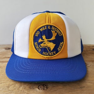 Puede incluir: Gorra de béisbol azul, blanca y dorada con visera plana. La gorra presenta un parche circular con un motivo de alce y el texto "100 Mile & District Minor Hockey Assn."