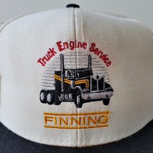 Op de afbeelding: Cr&egrave;mekleurige baseballpet met een zwarte klep. De pet heeft een geborduurd ontwerp van een zwart-gele vrachtwagen met de woorden "Truck Engine Service" in rood en "FINNING" in goud.