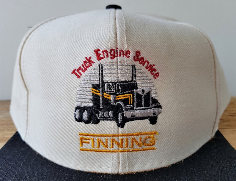 Op de afbeelding: Cr&egrave;mekleurige baseballpet met een zwarte klep. De pet heeft een geborduurd ontwerp van een zwart-gele vrachtwagen met de woorden "Truck Engine Service" in rood en "FINNING" in goud.